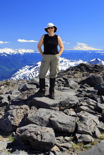 Female hiker wearing a sun hat, sleeveless black shirt and hiking pants, standing with her hands in her pockets atop a boulder field on a mountain slop