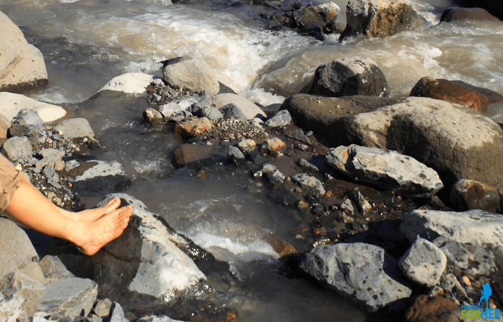 Feet of a hiker soaking in an icy glacier melt stream, with lots of boulders
