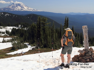 Female hiker standing on snow wearing yellow and gray backpack with trekking poles planted  near a hiking trail sign with Mount Adams in the distance