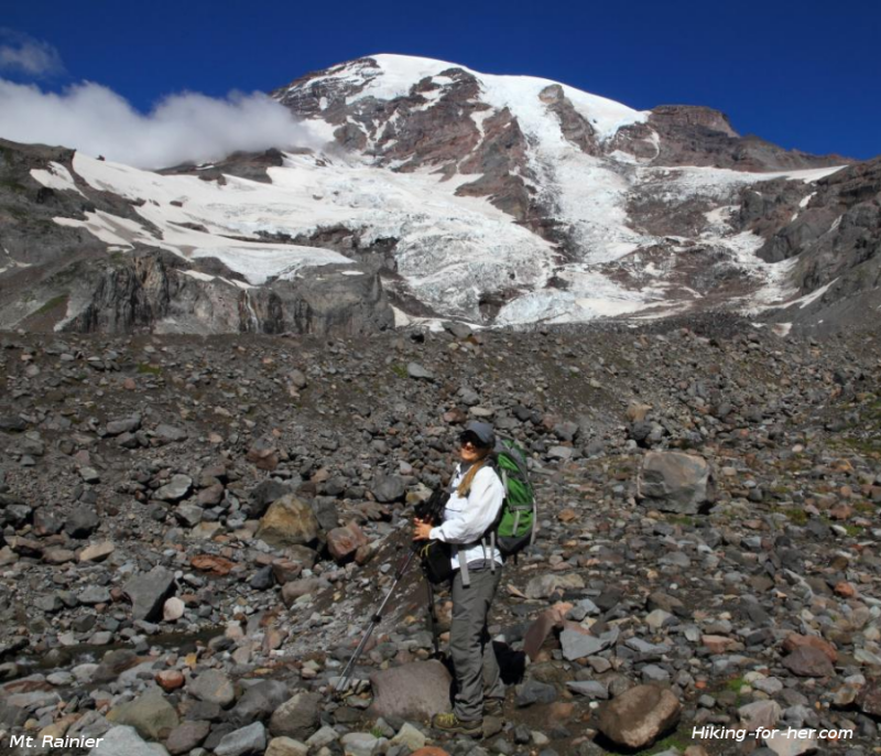 Female hiker on rocky slope with snow and ice covered Mount Rainier and blue sky in background Female hiker on rocky slope with snow and ice covered Mount Rainier and blue sky in background