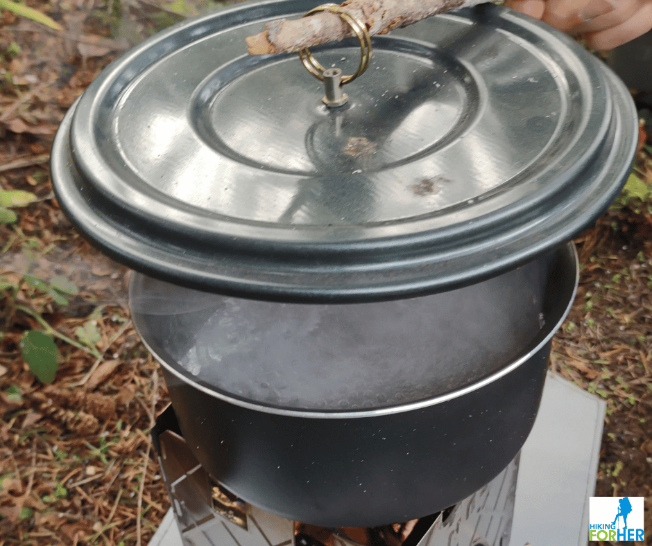 Ganesha ultralight backpacking twig stove showing a pot filled with boiling water as a small branch is used to lift the lid off the pot