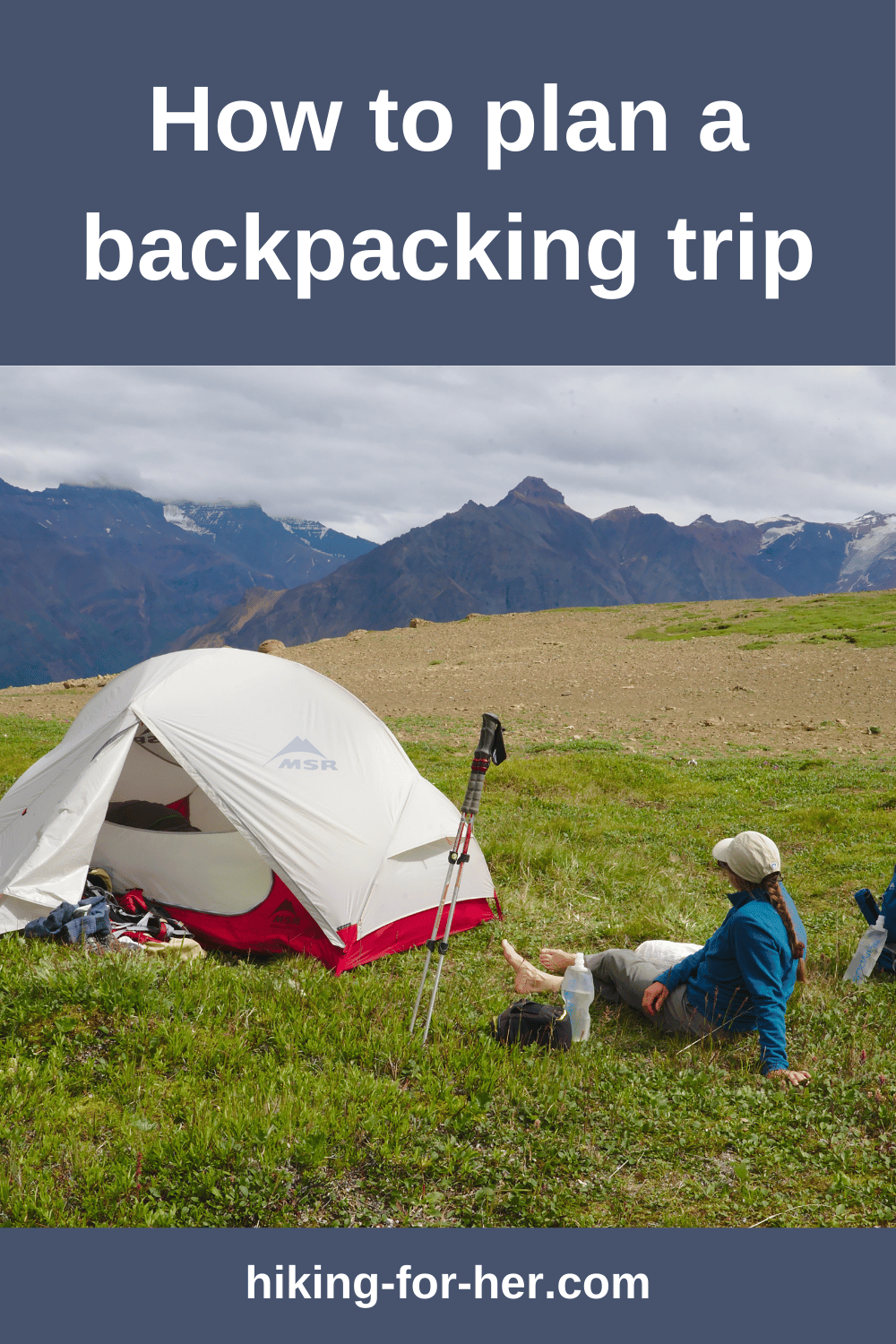female hiker sitting with boots off outside a red and white tent and hiking gear with mountains in background