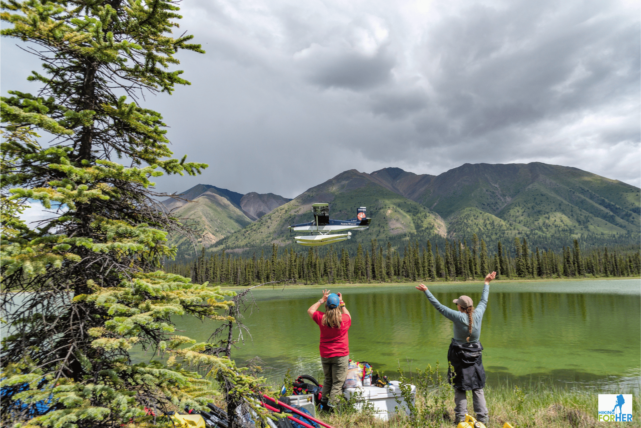 Two female hikers at alpine lake waving goodbye to bush plane on floats taking off