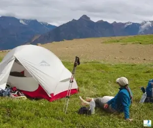 Female backpacker sitting beside tent and backpack with mountains in background Female backpacker sitting beside tent and backpack with mountains in background
